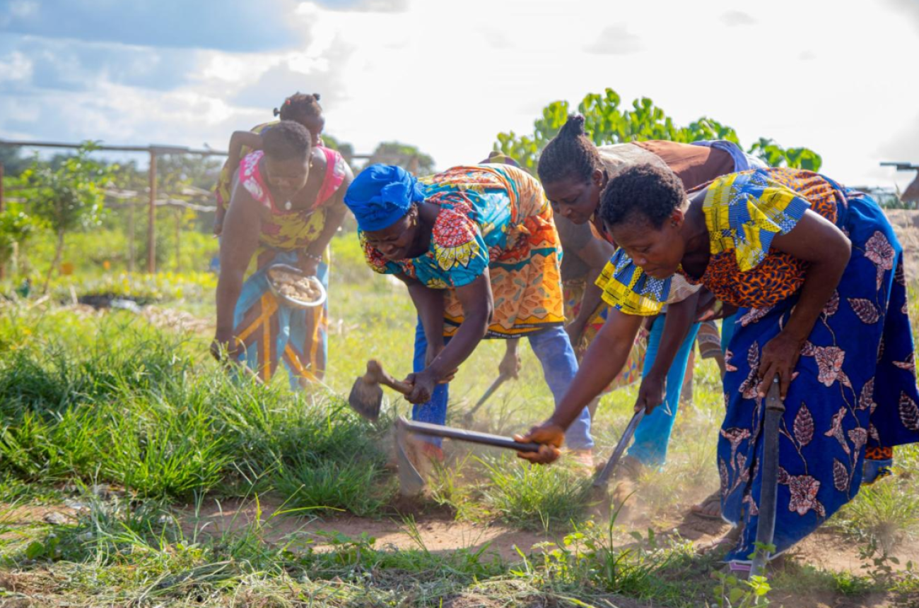 Mujeres con faldas y tops de colores labran la tierra en medio de un paraje lleno de vegetación.