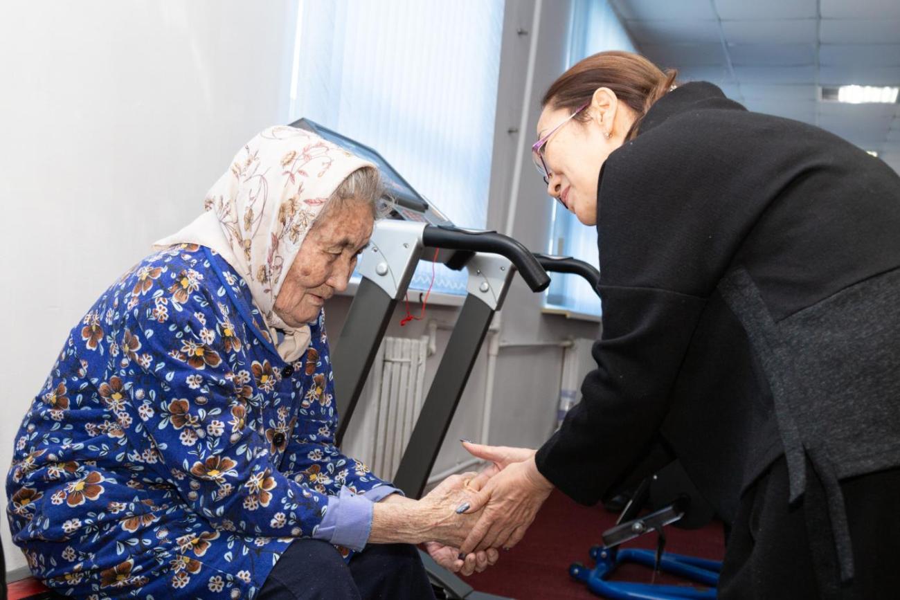 Older woman on the left in blue dress and headscarf holding hands with woman on the right in a black dress