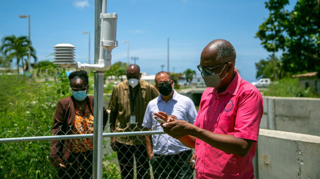 A man in a pink shirt in the foreground analyzes data on a small electronic device, while other officials look on.