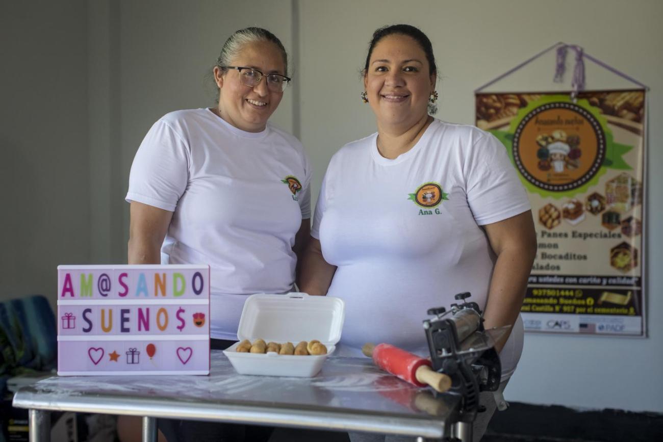 Two women in white shirts standing in a bakery kitchen with kitchen tools and baked goods