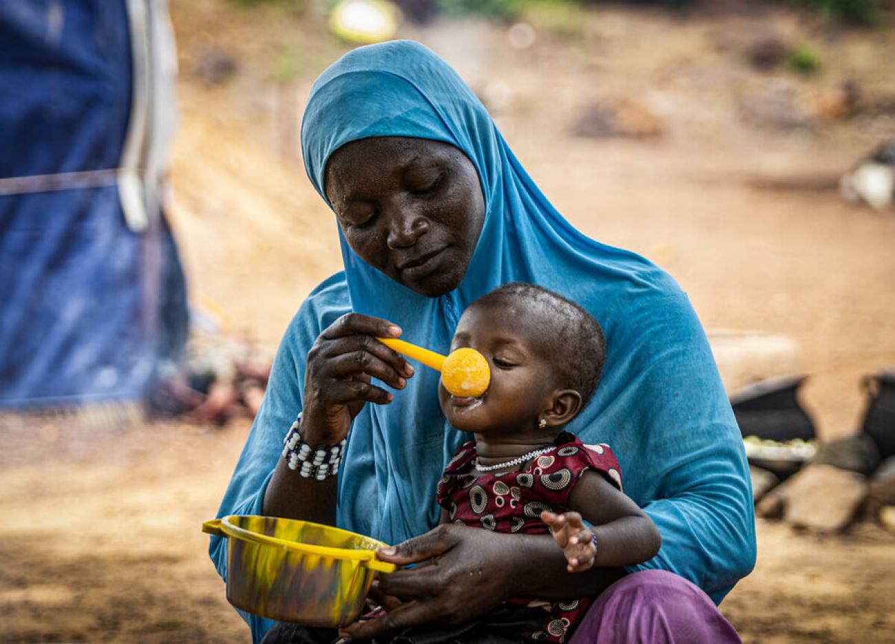 Woman in a blue dress and headscarf feeds a baby with a steel bowl and a yellow spoon