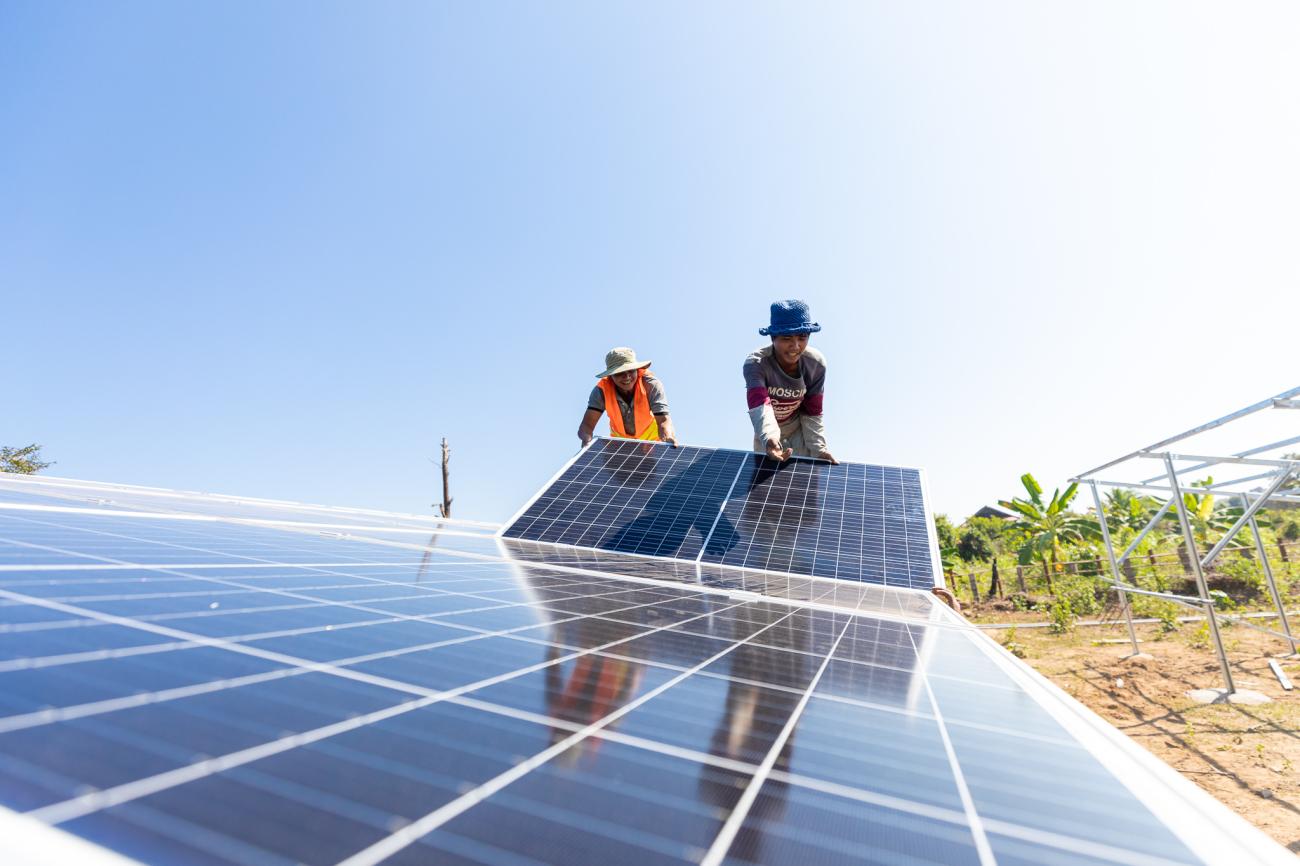 Two people in hats and colourful clothes stand on one side of a silver coloured solar panel in Cambodia