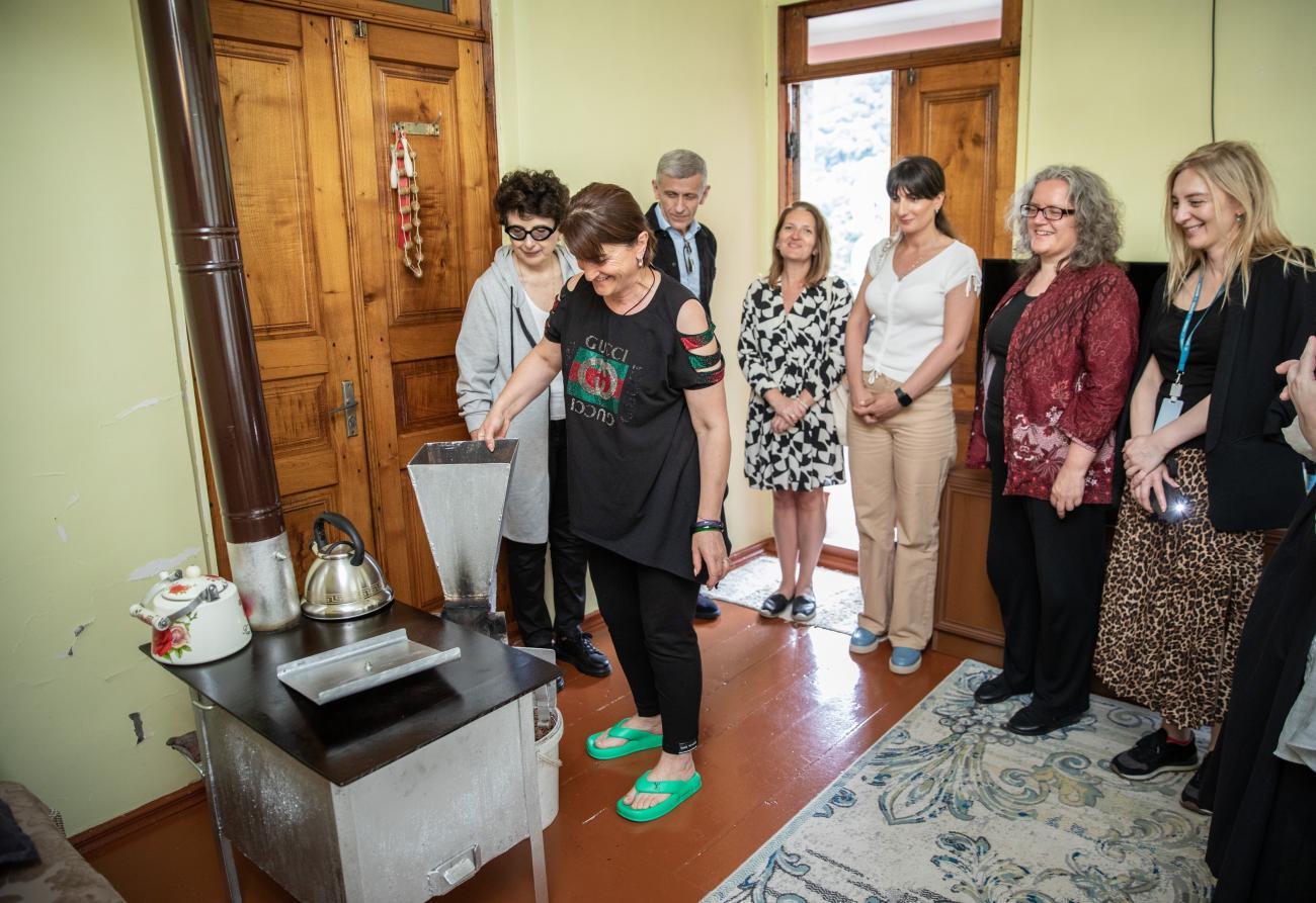 A group of people gather around a woman in a black shirt inside a house. The woman is putting shells into a silver heating machine.