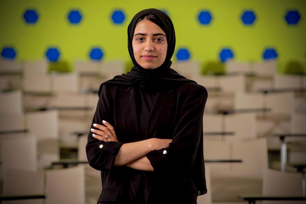 An Afghan girl in a black dress and headscarf stands in front of a polka dot wall