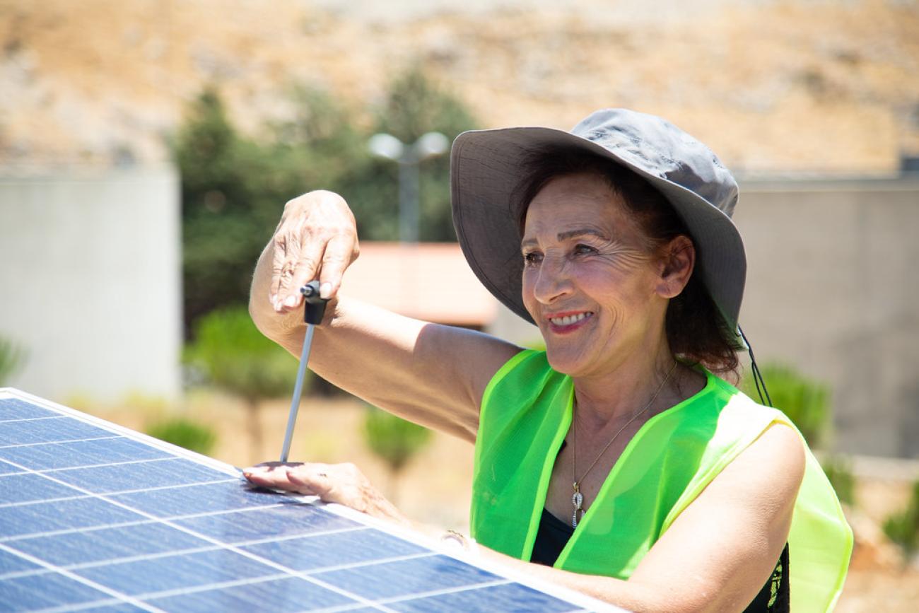 A woman in a hat and a green vest works with a tool fixing a solar panel