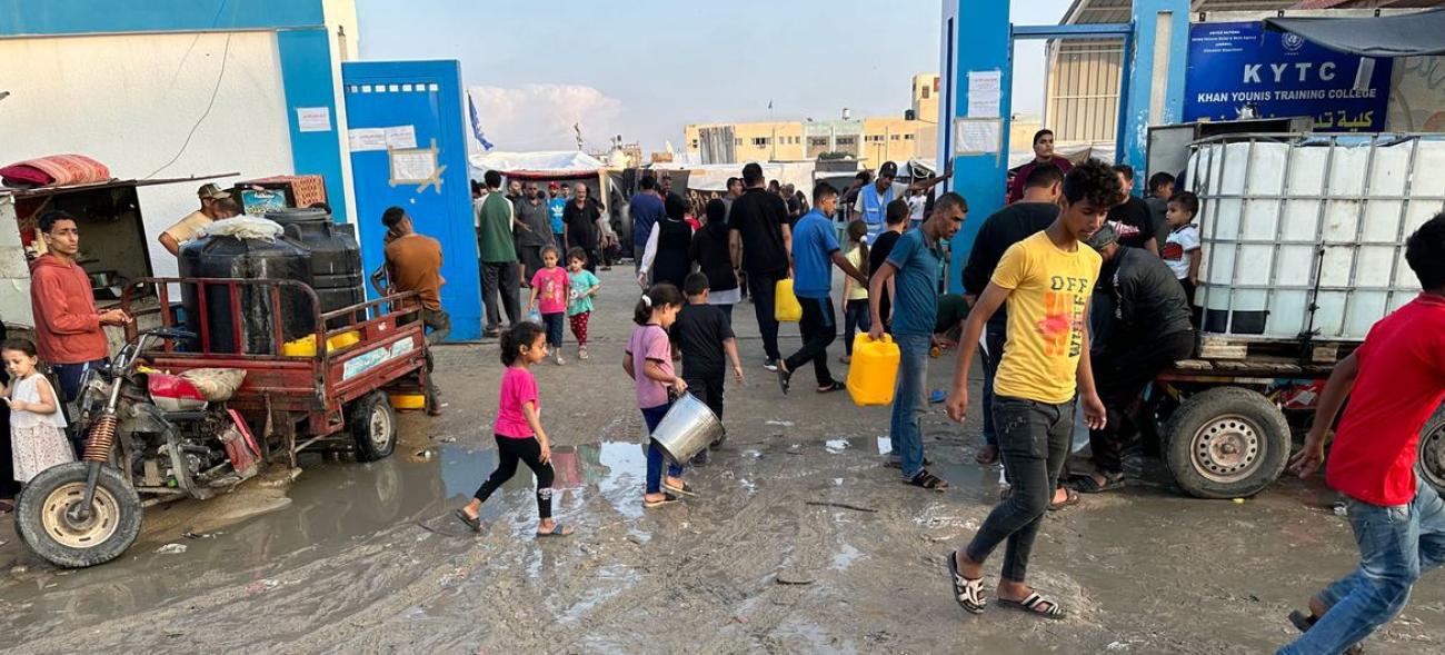 People walk around what looks like a crowded market on muddy ground. 