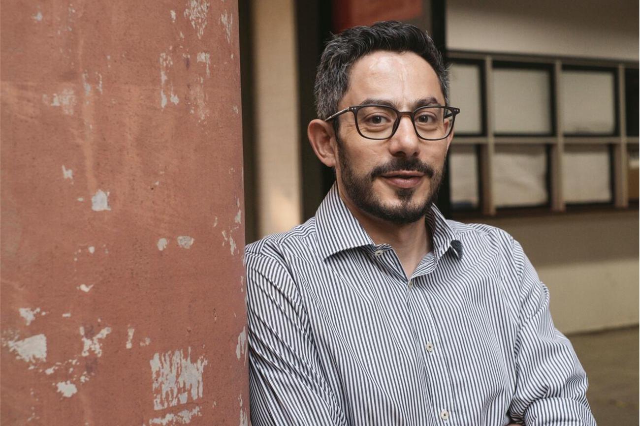 A man in a grey shirt and glasses stands next to a wall painted brown.