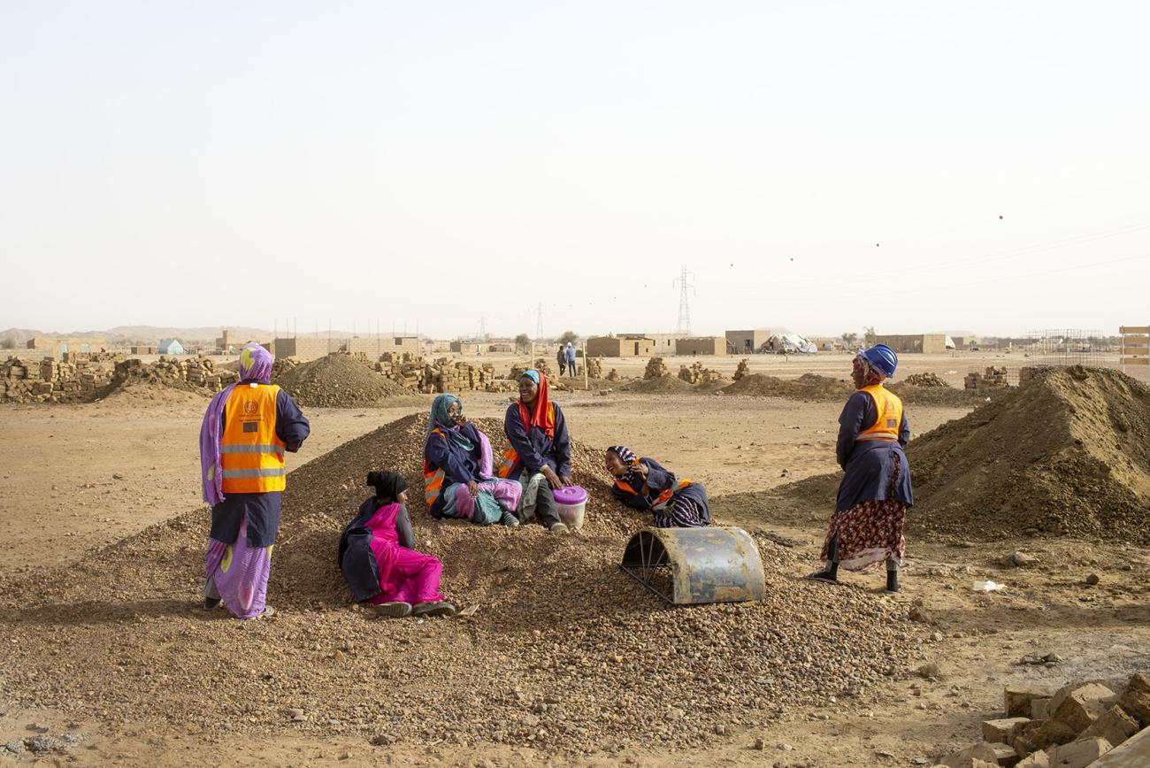 Women in colorful dress and hard hats sit and stand at a small construction site with holes in the ground.