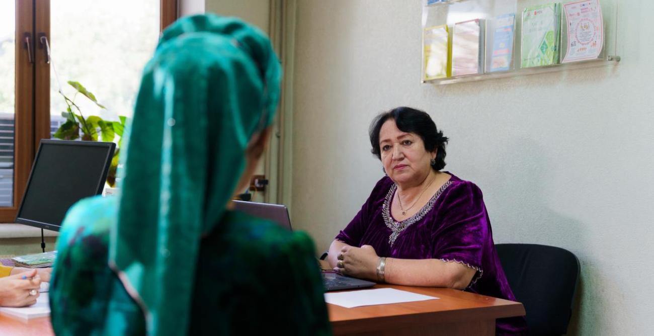 A woman in a green dress sits in a chair and talks to another woman in a purple dress in Tajikistan.