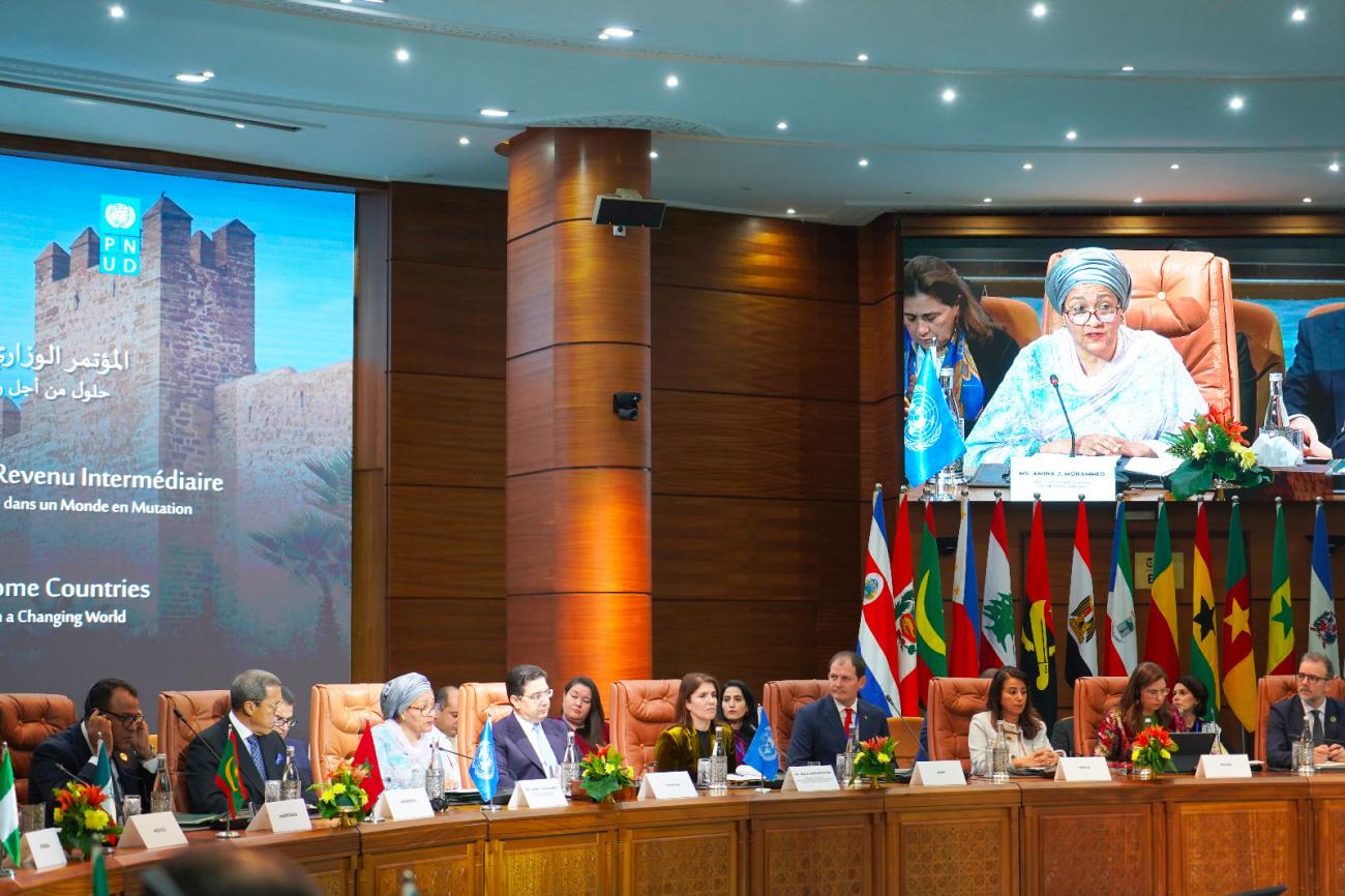 The deputy head of the UN sits on a panel with dignitaries and appears on a wide screen, with country's flags and flowers around the table.