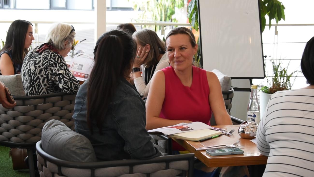 A woman in a red shirt talks to another woman in a black dress with other women in the background sitting around a table