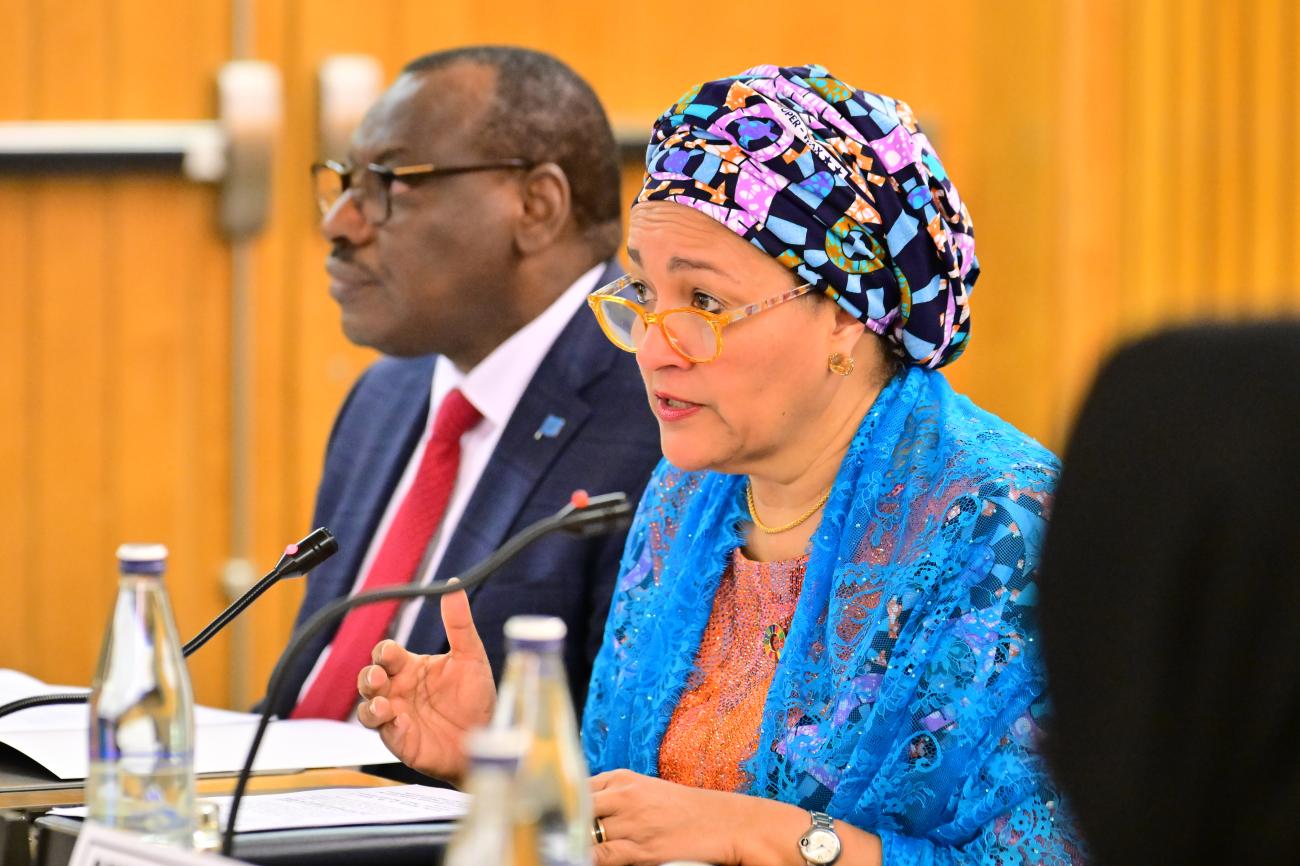 A woman in a blue dress and colourful headscarf, the UN Deputy Chief Amina J. Mohammed, speaks into a microphone at a podium. Another man in a blue suit and a red tie sits to her right side.