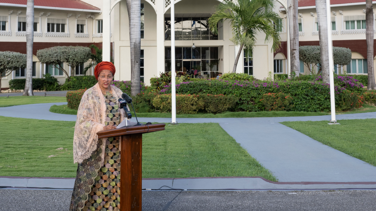 A woman standing at a podium with a microphone, set up outdoors in front of a building with white columns and multiple stories.