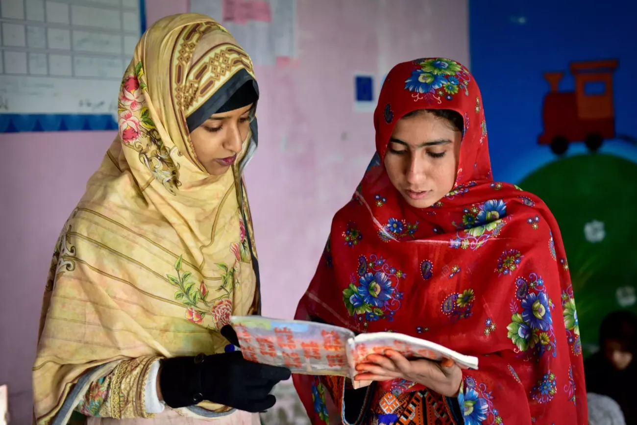 A girl in a yellow headscarf and dress reads a book with another girl in a red headscarf and dress inside a classroom