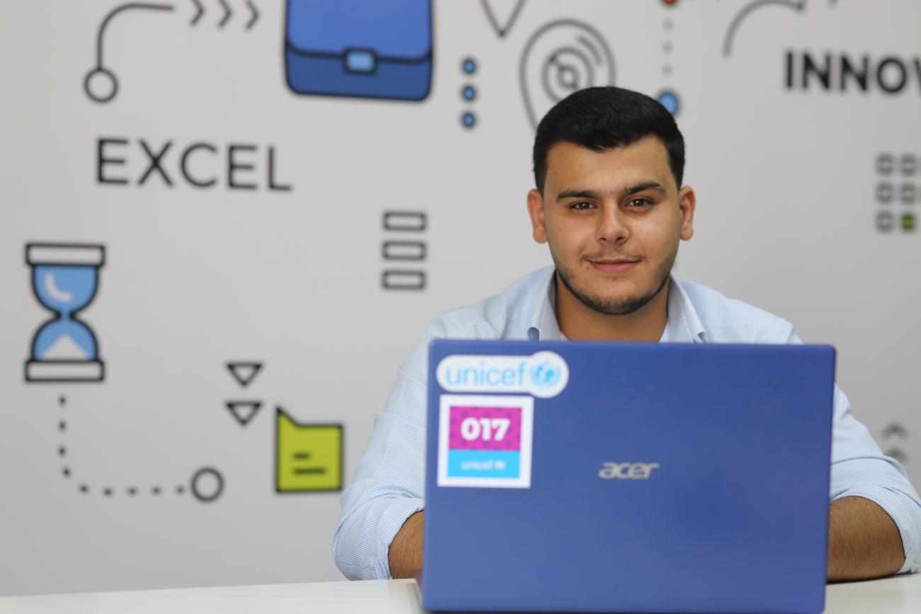 A boy is seated behind a laptop and looking at the camera. He has a colourful background behind him