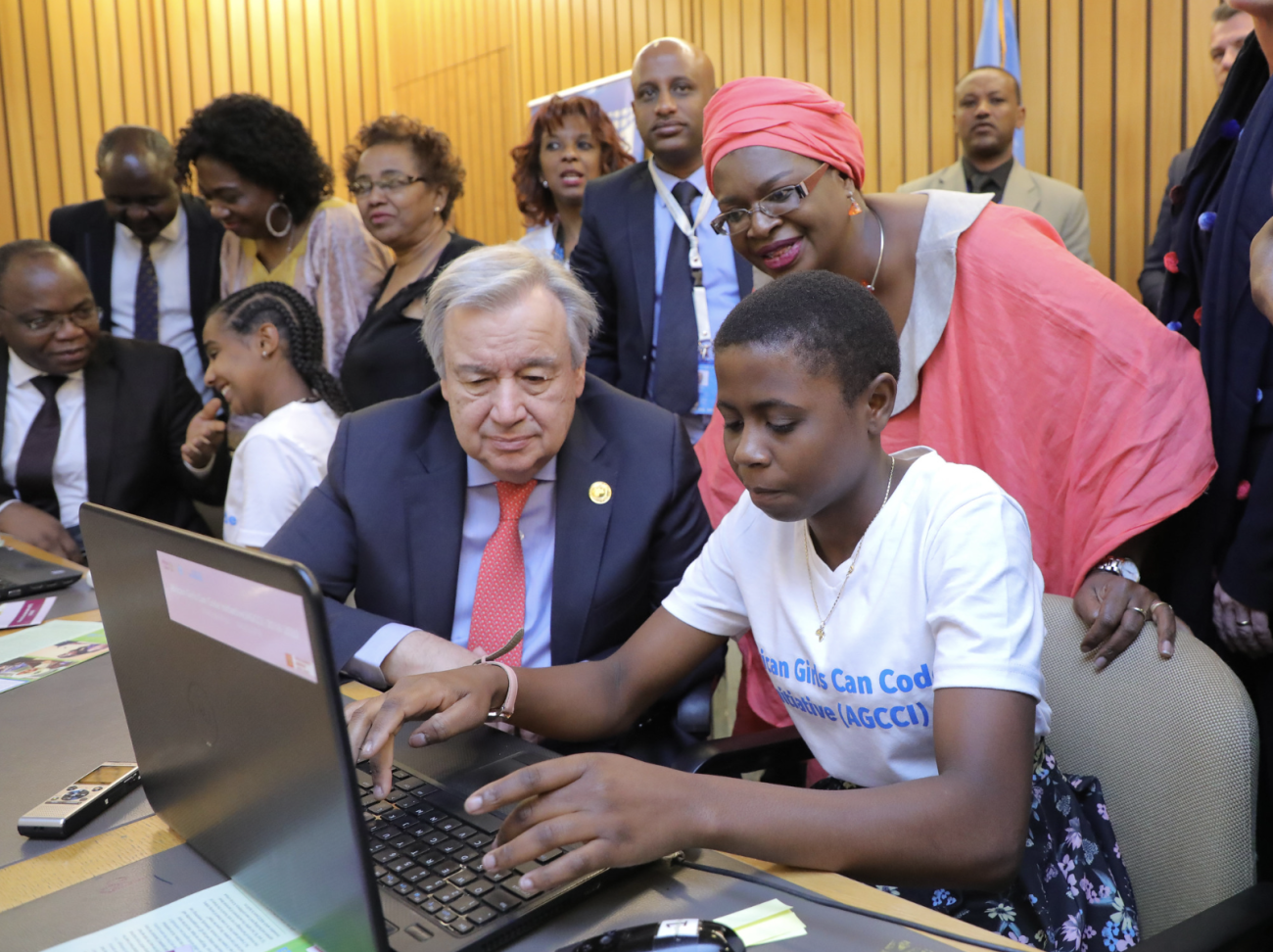 A group of young people along with the Secretary-General Antonio Guterres, peek into a computer as one of them shows them the screen.