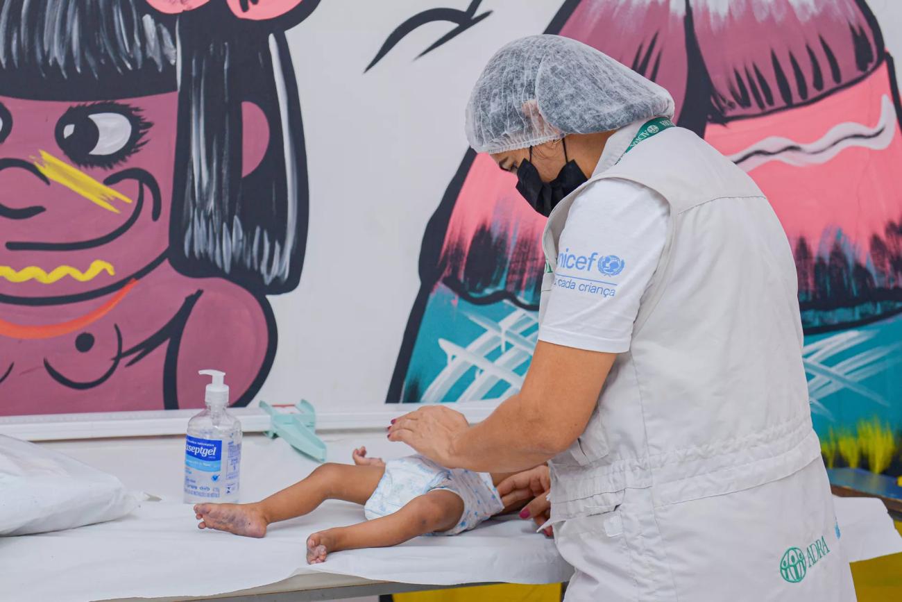 A woman in a grey vest and headcap looks over a baby on a clinic bed.