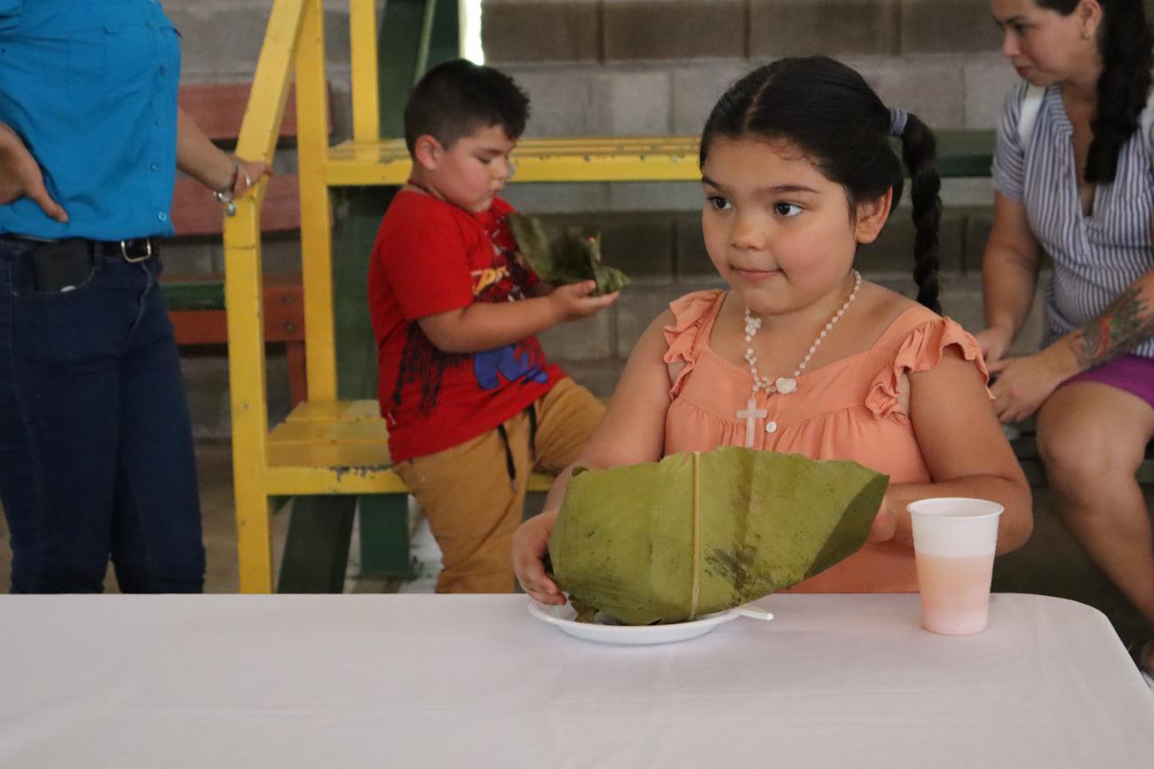 A little girl in an orange shirt sits at a table with a plate of tamales wrapped in leaves and a cup of liquid next to it. Behind her in a boy in a red shirt.