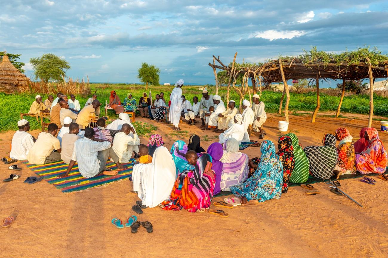 A panoramic view of a group of community members gathered. The women sit across from the men, wearing colourful full length clothes, wile the men sit on the other side dressed in white and with head-gears.