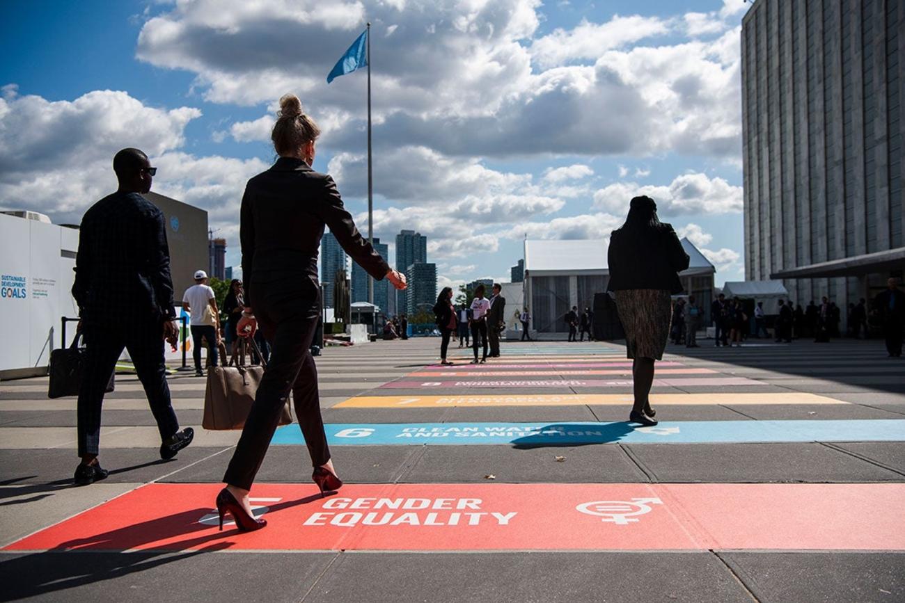 Silhouettes of three people walk through an open lobby at the UN in New York as the ground carries a painted sign of SDG Goal 5 on gender equality
