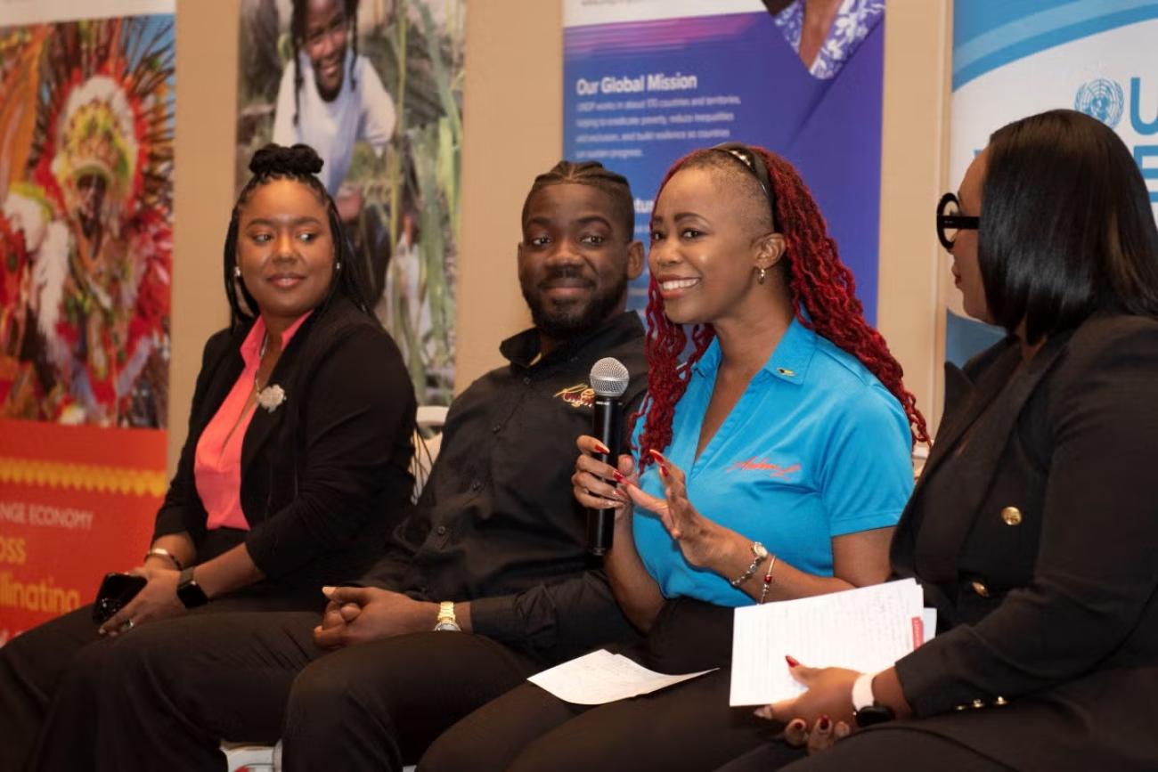 A woman in a blue t-shirt speaks into a microphone while three other people, two women and a man listen intently to her.