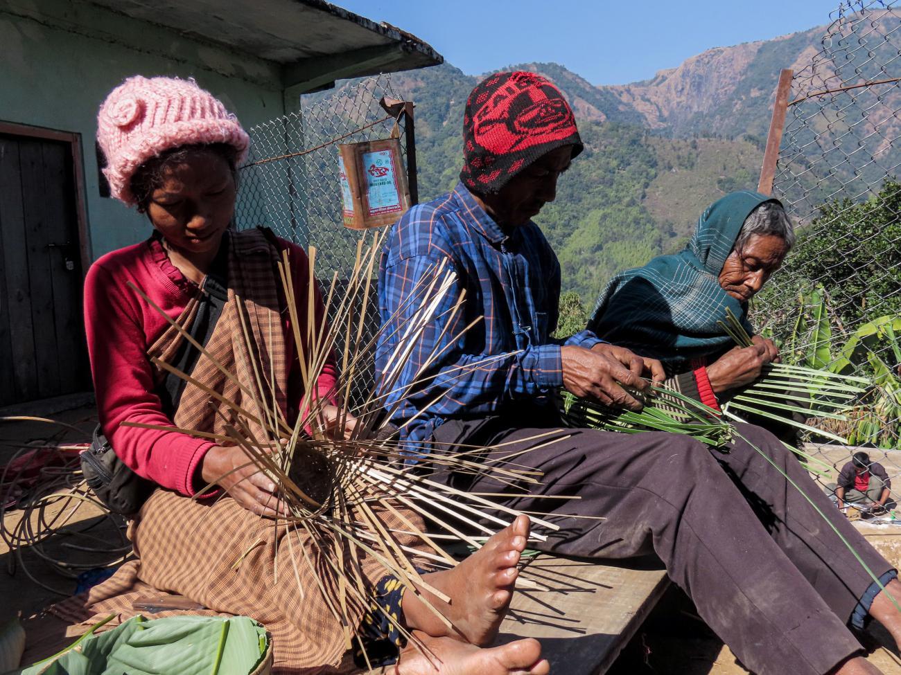 A woman and two men are sitting together weaving bamboo strands.