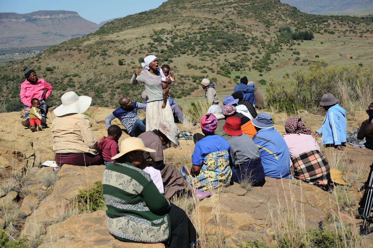 A woman stands in the middle of a group outdoors and speaks to them, while holding her child