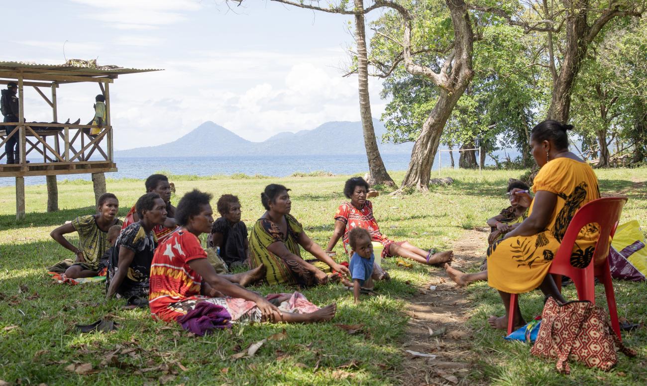A group of people are gathered sitting on the ground, in front of a woman sitting on a chair talking to them