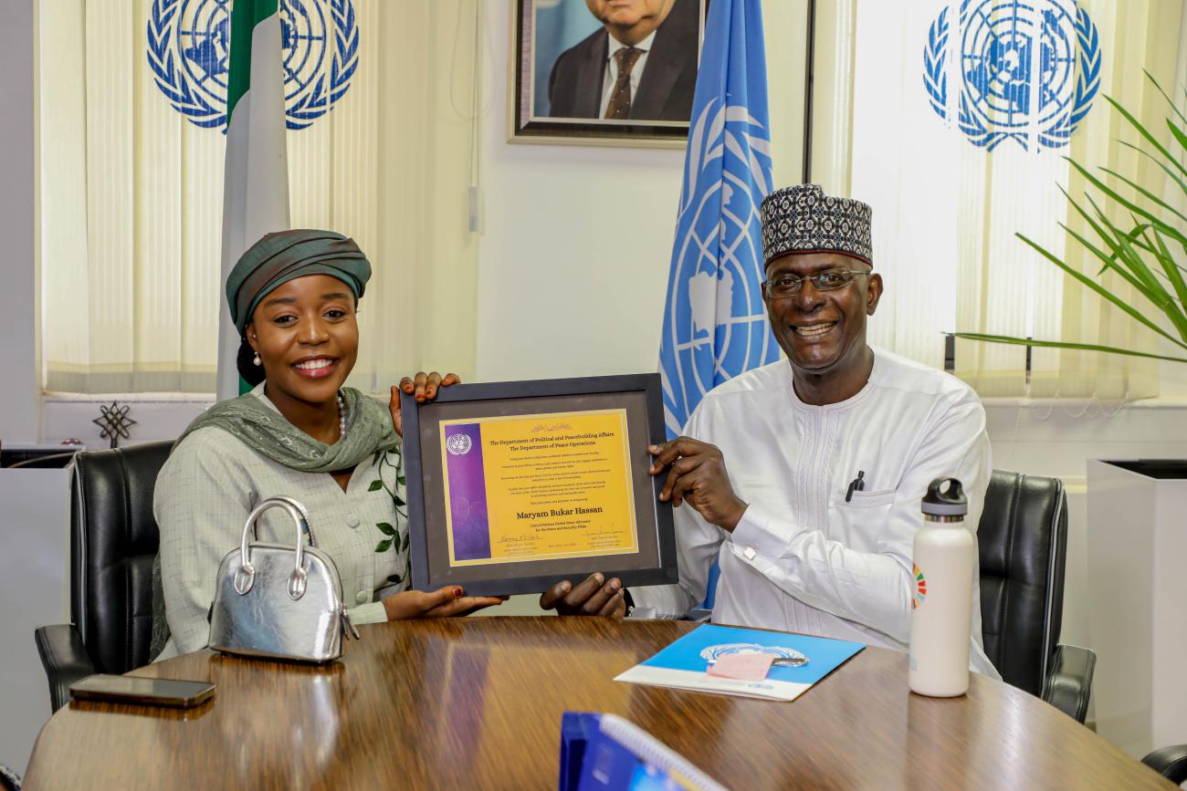A man in white clothing hands over a commemorative plaque to a woman in a green dress and headdress