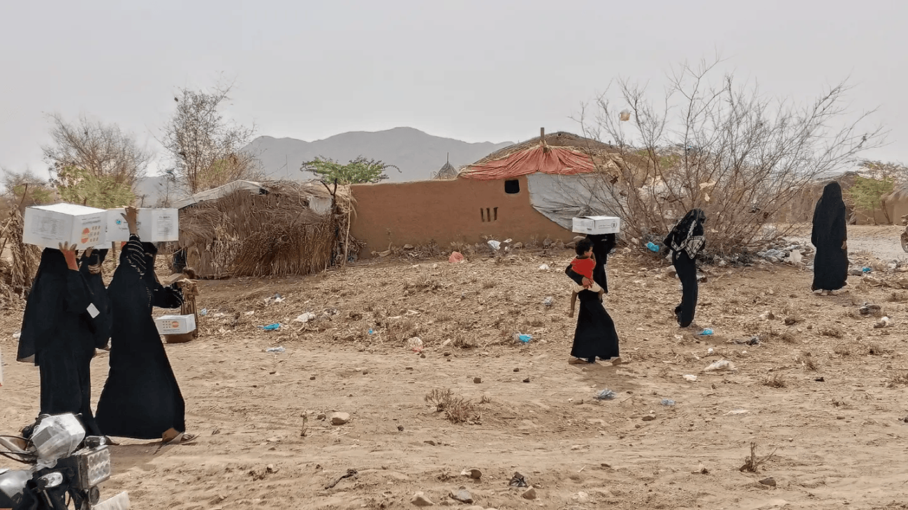 نساء وأطفال في مديرية عبس بمحافظة حجة يحملون صناديق مساعدات غذائية أمام بيوت طينية وأكواخ بسيطة، في مشهد يجسّد معاناة الأسر مع تفاقم الجوع.