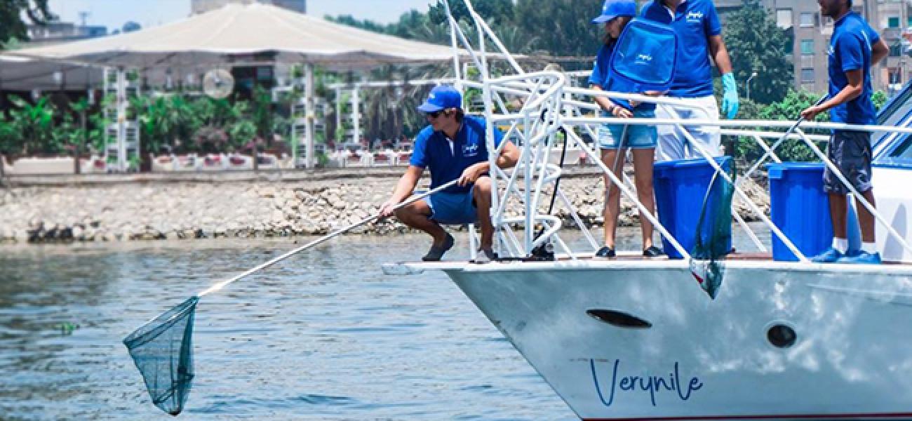 À bord d’un bateau de taille moyenne baptisé Verynile, des personnes portant un t-shirt bleu, une casquette bleue et un short s’emploient à nettoyer les déchets déversés dans le Nil. Parmi elles, un homme, accroupi, tend une longue épuisette à manche vers l’eau du fleuve.