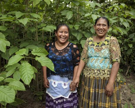 En medio de un área verde (vegetación tropical y húmeda), dos mujeres indígenas sonríen a la cámara.