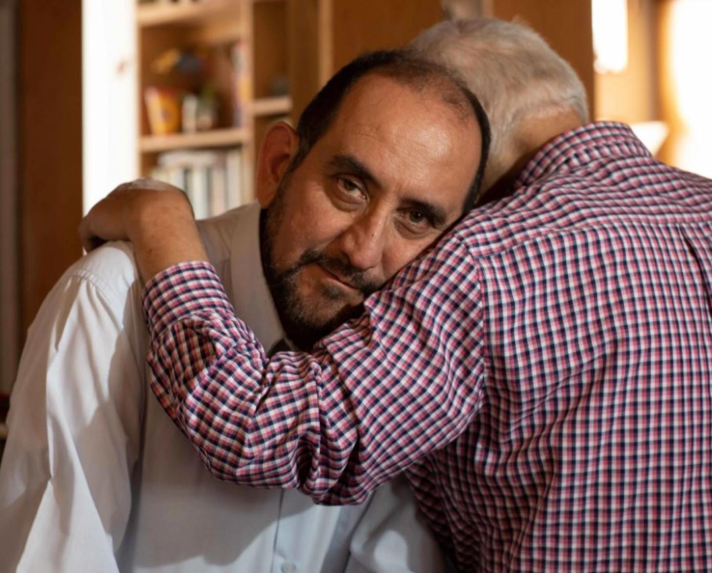 Alejandro mira a la cámara mientras Camilo se sienta frente a él de espaldas a la cámara y con su brazo rodeando cariñosamente a Alejandro.