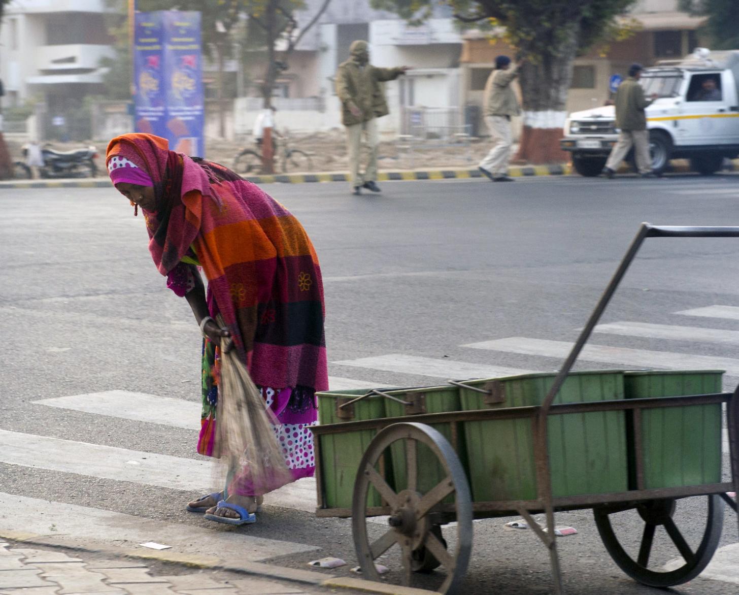 Una mujer ataviada con un velo que cubre la cabeza, el pecho y le llega hasta las rodillas, tira de una carrucha verde sobre un paso de peatones, en la calle.