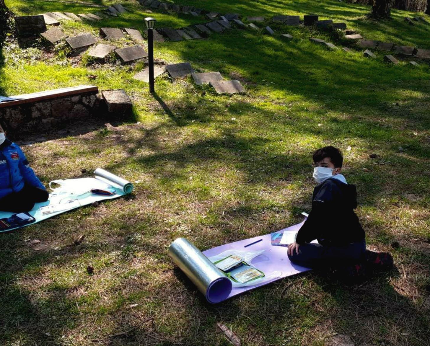 Young students wearing protective gear take their online courses outside.