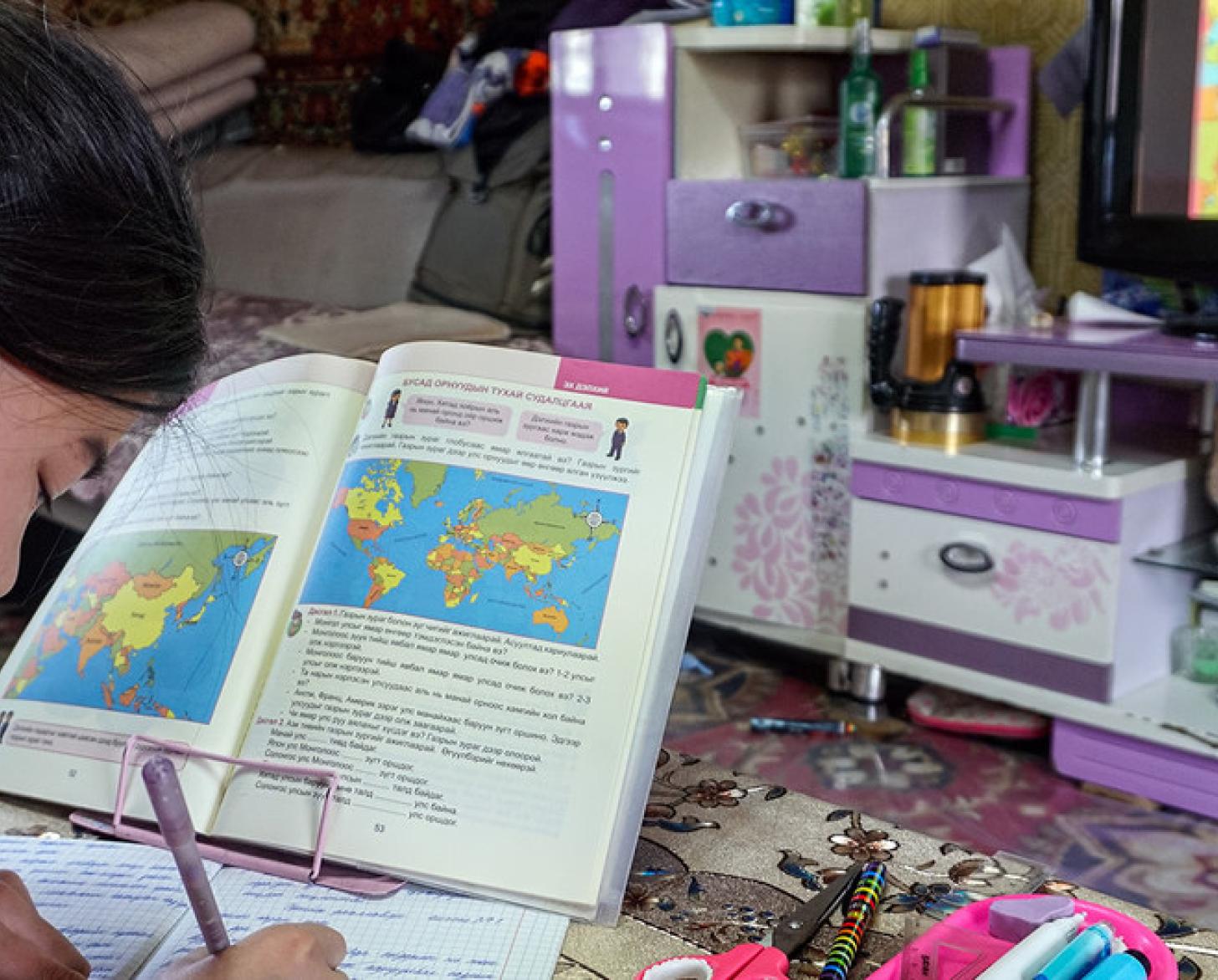 Une petite fille fait consciencieusement ses devoirs, assise à une table où sont disposés un livre de géographie et des jouets.