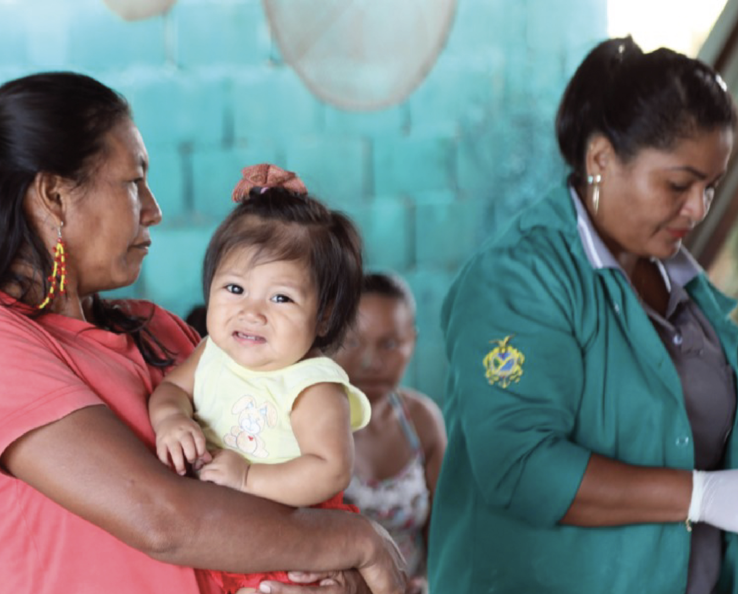 Une femme autochtone tient une petite fille dans les bras, à gauche de l’image, en attendant d'être soignée par une agente de santé dont on voit qu’elle prépare son matériel à droite de l’image. La femme autochtone regarde en direction de l’agente de santé tandis que sa fille regarde la caméra le visage grimaçant.