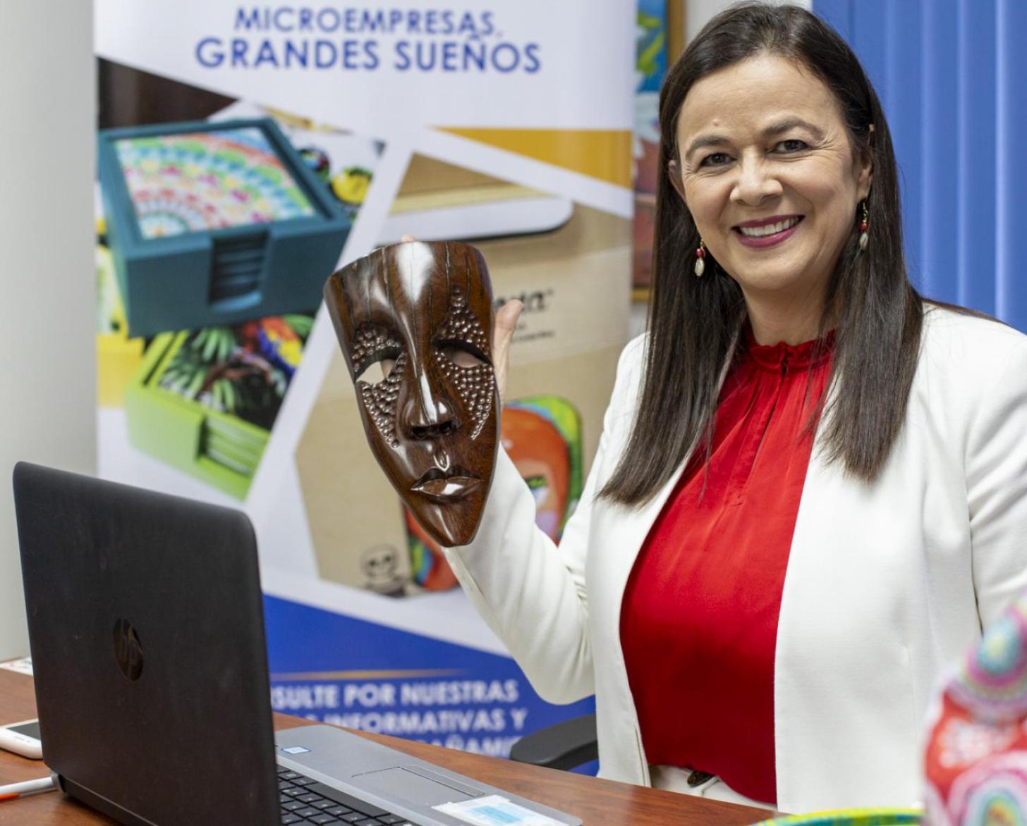 A woman smiles happily as she stands at a desk holding a mask.