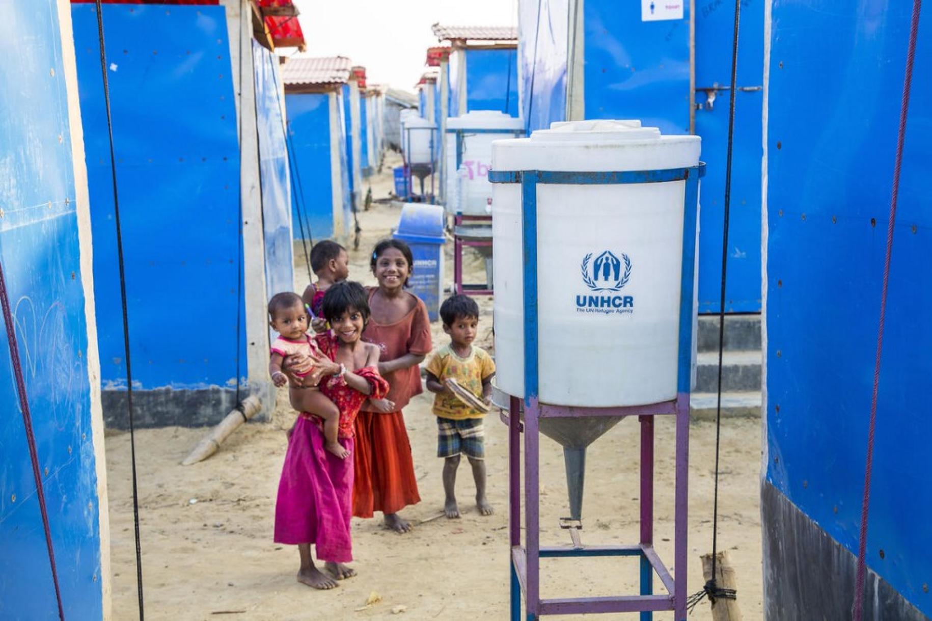 Des enfants se tiennent souriants, face à la caméra, près d'une installation pour le lavage des mains du camp de réfugiés de Kutupalong, au Bangladesh.