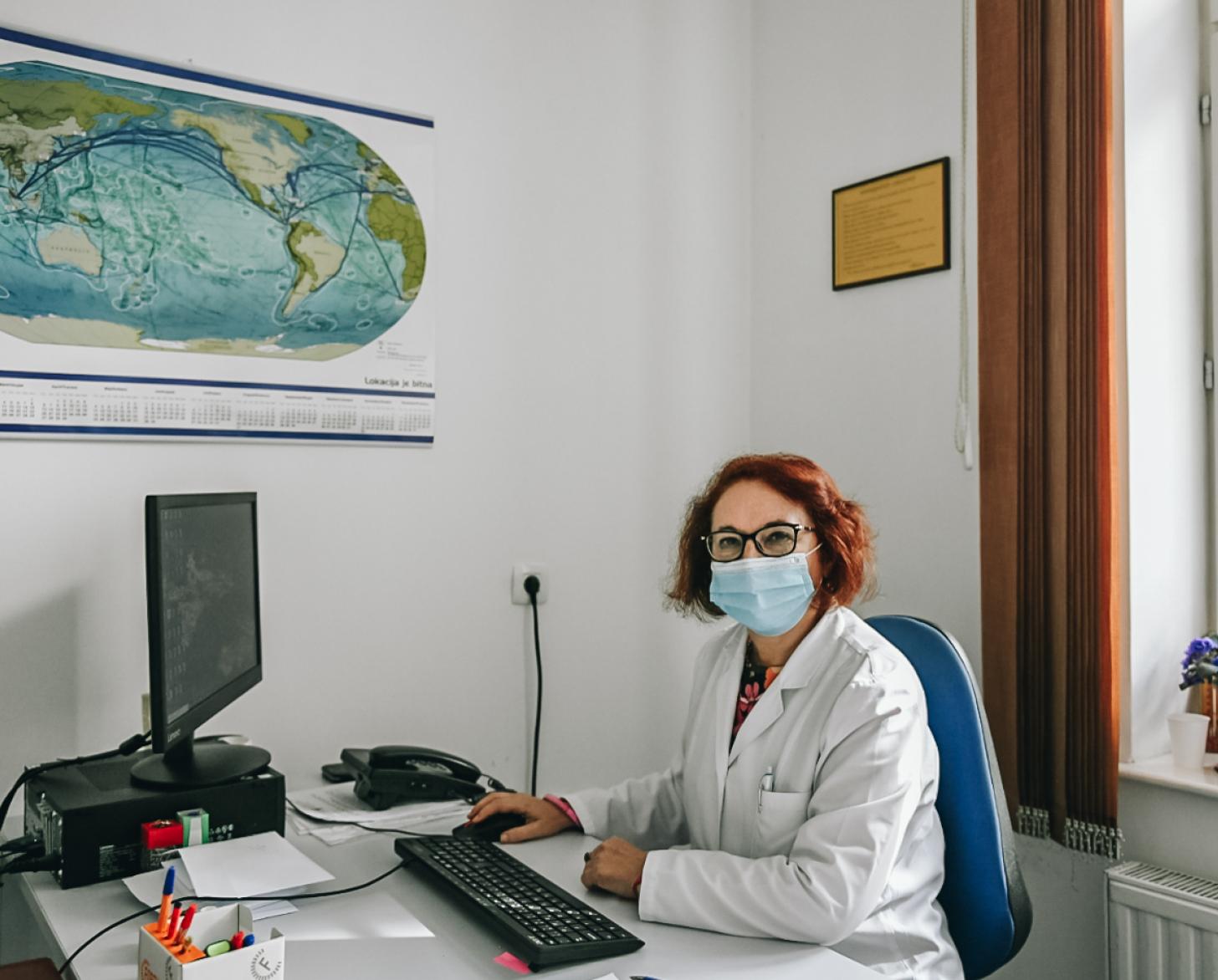Dr. Snežana sitting at her desk with a poster of a world map just above her desk.