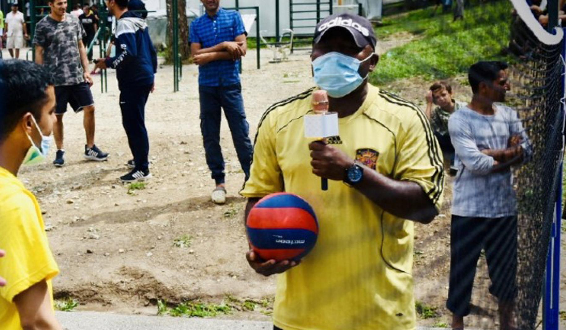 Jean Marc Bogmis sostiene un micrófono en una mano y un balón en la otra mientras la gente le rodea en un terreno descampado.