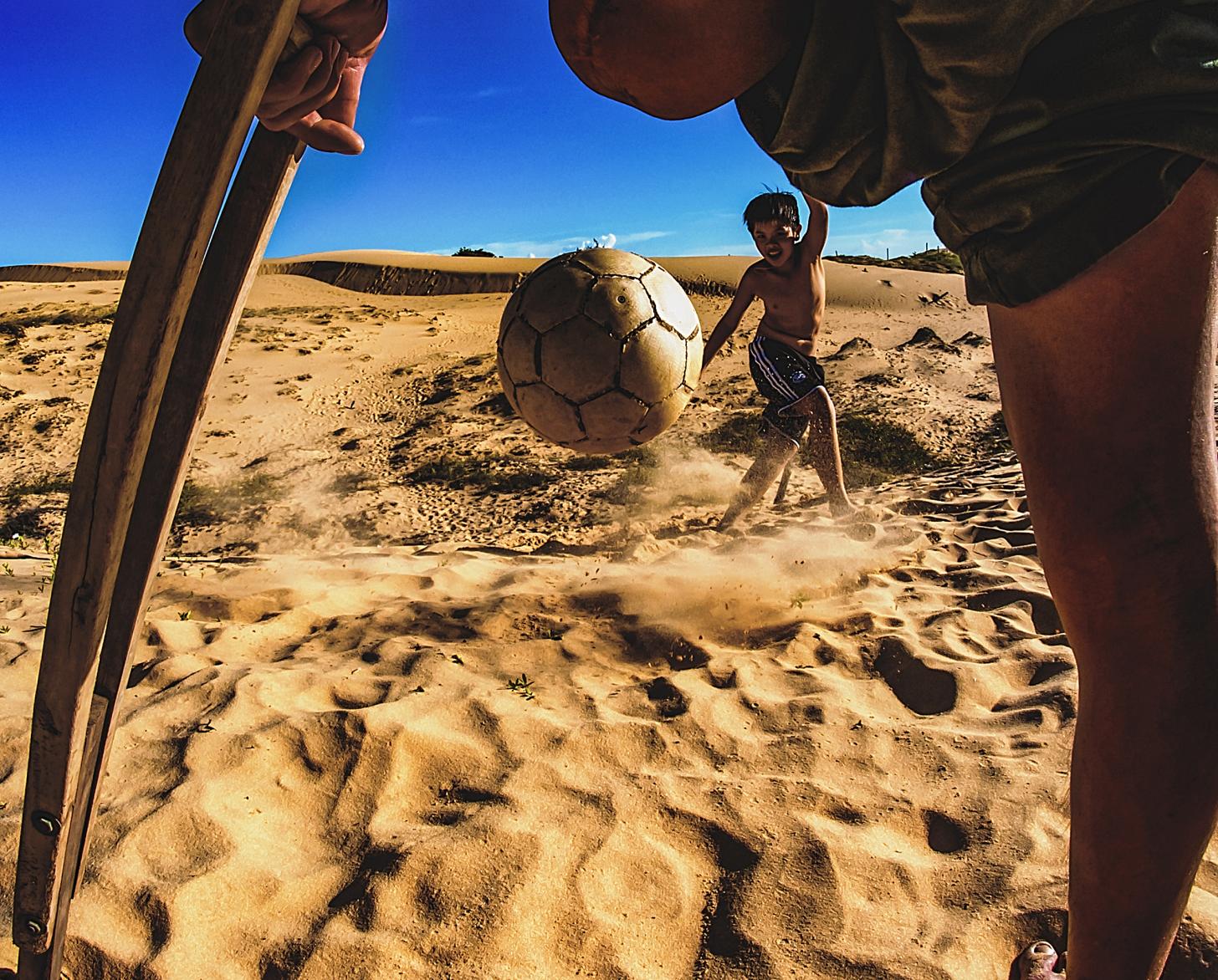 Photo shows a close-up between the legs of an man with an amputated leg. Through this angle you can see a soccer ball being kicked towards the man by a young boy.