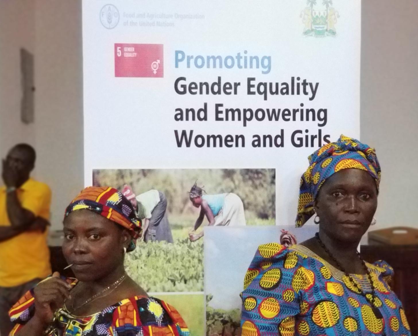 Two women stand powerfully in front of an banner promoting SDG 5: Gender equality and empowering women and girls.