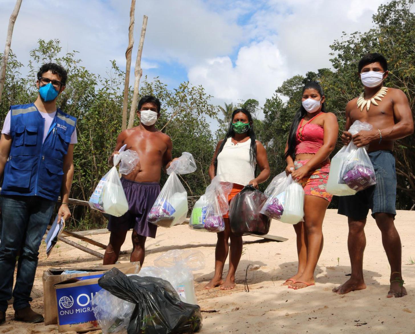 Un miembro del personal de la OIM y una familia indígena tras recibir los suministros, mantiene el distanciamiento social, mientras miran a la cámara (son cinco personas adultas, usando mascarillas)).