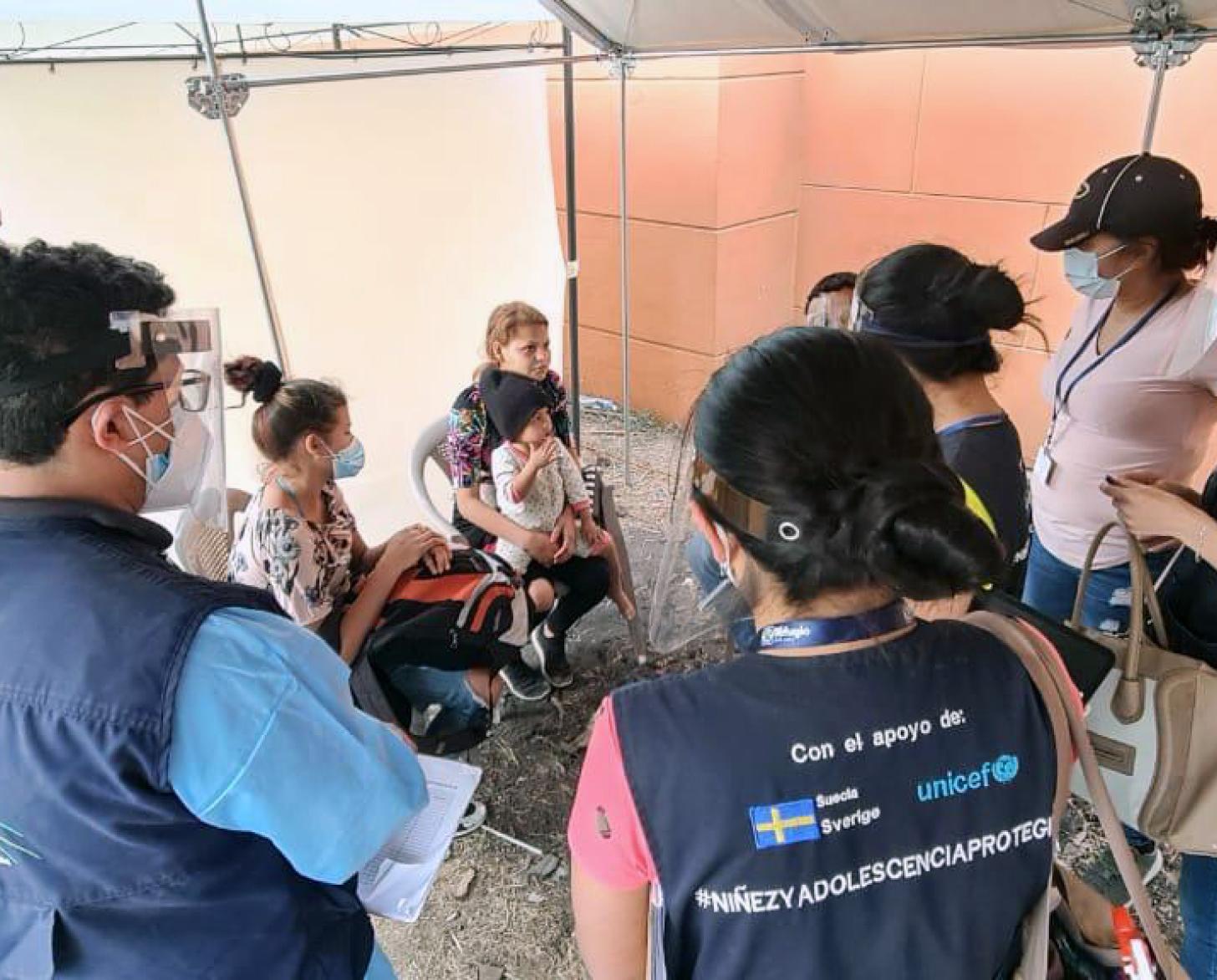 Personal de la ONU con mascarillas y protectores faciales rodean a una familia sentada en sillas en un círculo, todos se encuentran bajo una tienda de campaña.