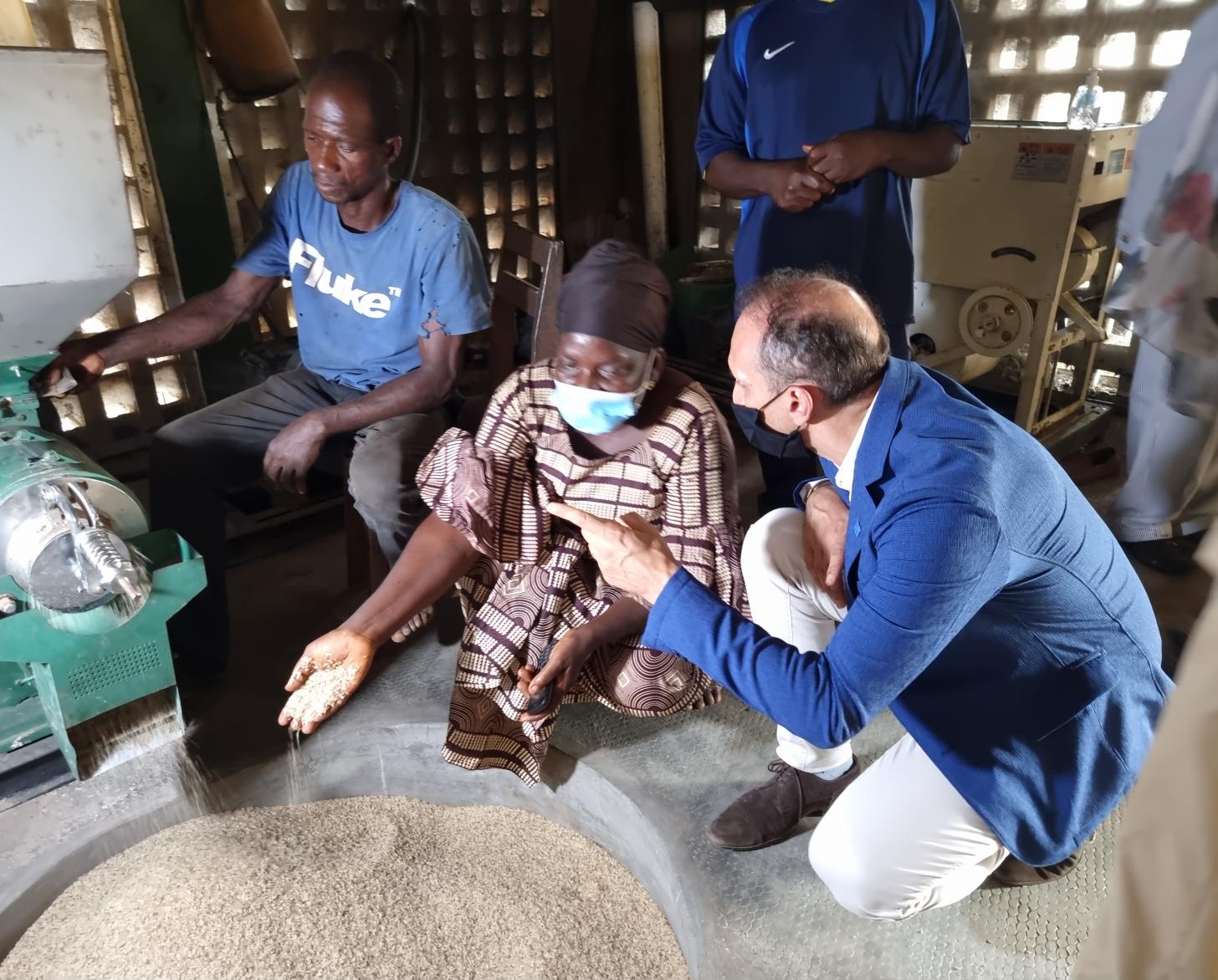 Le Coordonnateur résident Vincent Martin se tient aux côtés d’un leader de la communauté. Tous deux discutent, agenouillés, devant une machine qui déverse des grains dans un grand récipient rond.