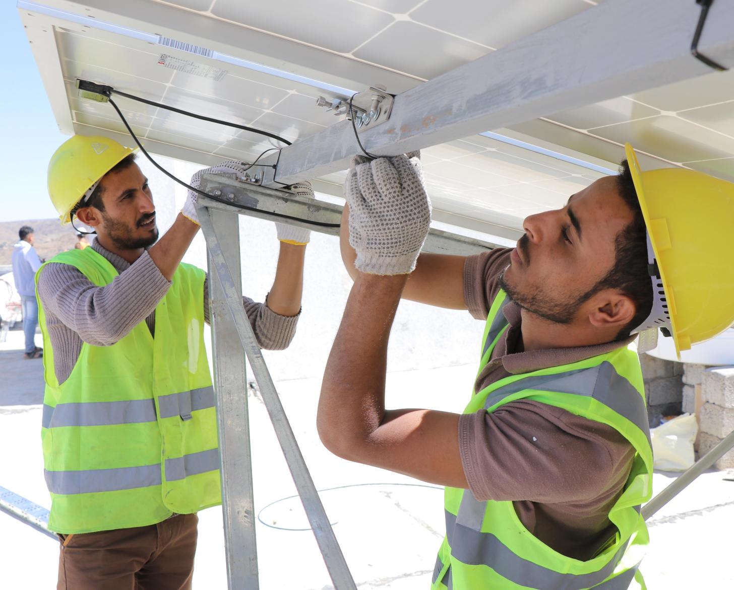Two men wearing hard hats and reflective jackets are working on solar panels.