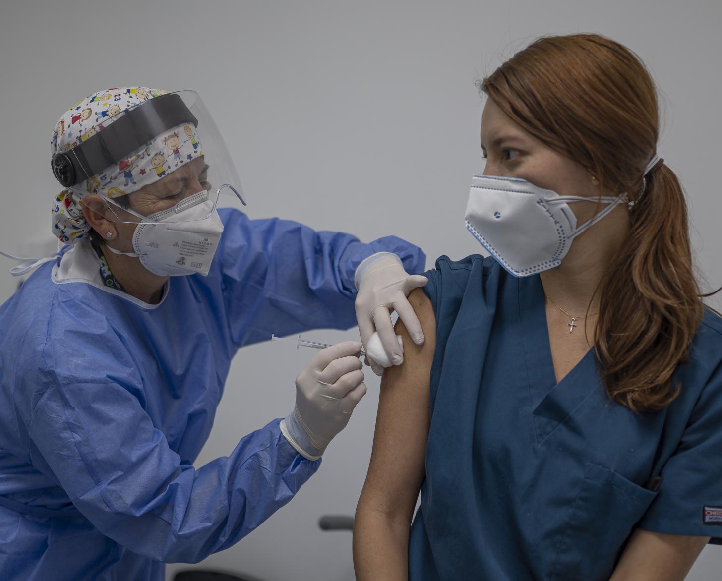 A healthcare professional wearing full head-to-toe protective gear is shown vaccinating a female colleague. 