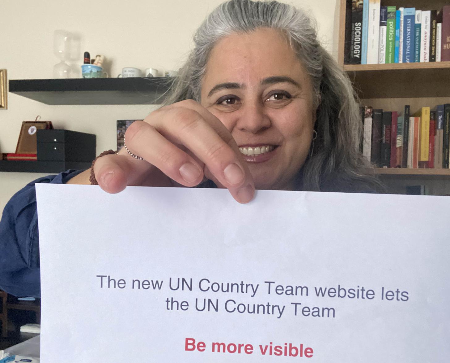 Una mujer sonríe alegremente a la cámara mientras sostiene un letrero que dice: "Los nuevos sitios web del equipo de país de la ONU permiten que el equipo de país de la ONU sea MÁS VISIBLE".