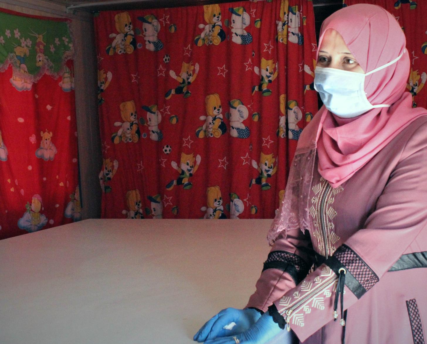 Haifa, 33, wears a face mask and gloves as she stands by a table in a room with red curtains.